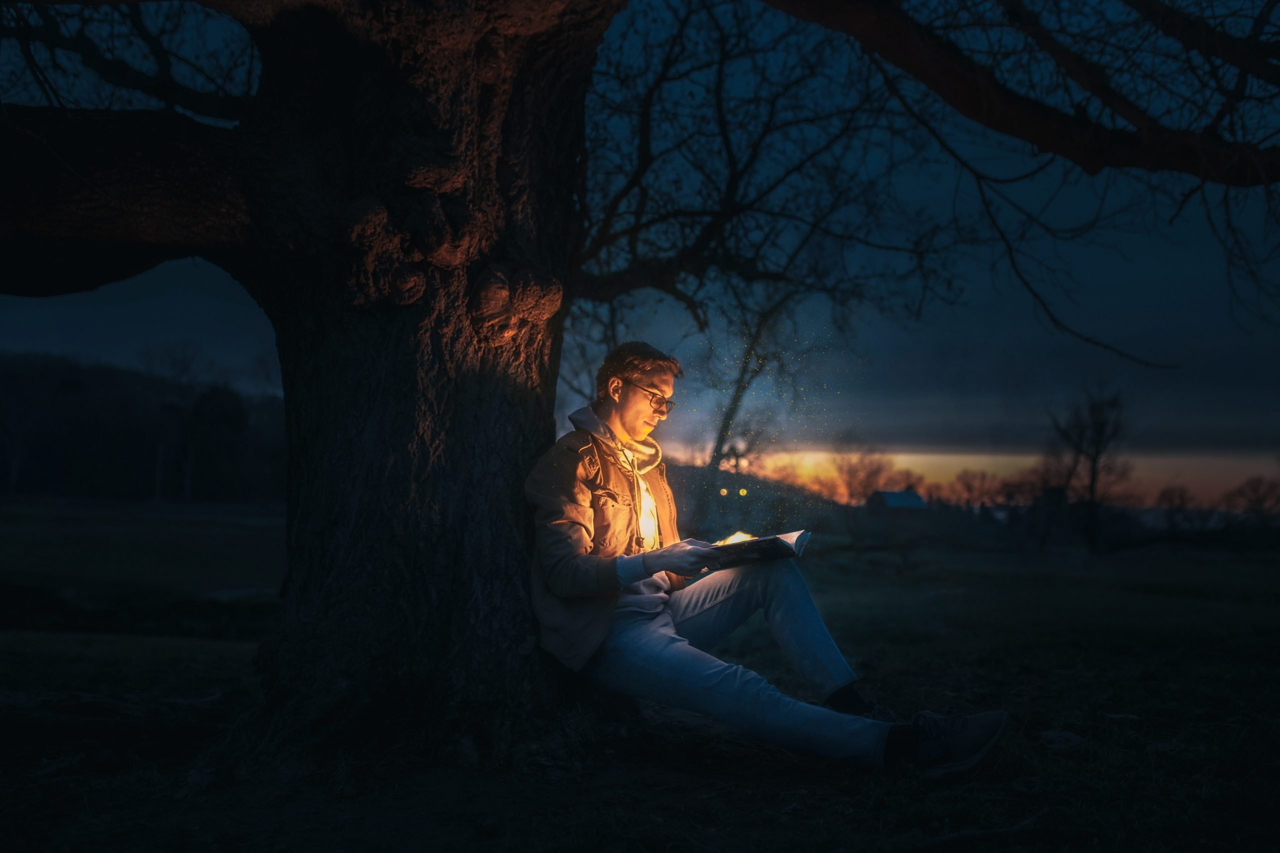 young man sitting at base of tree holding glowing open book